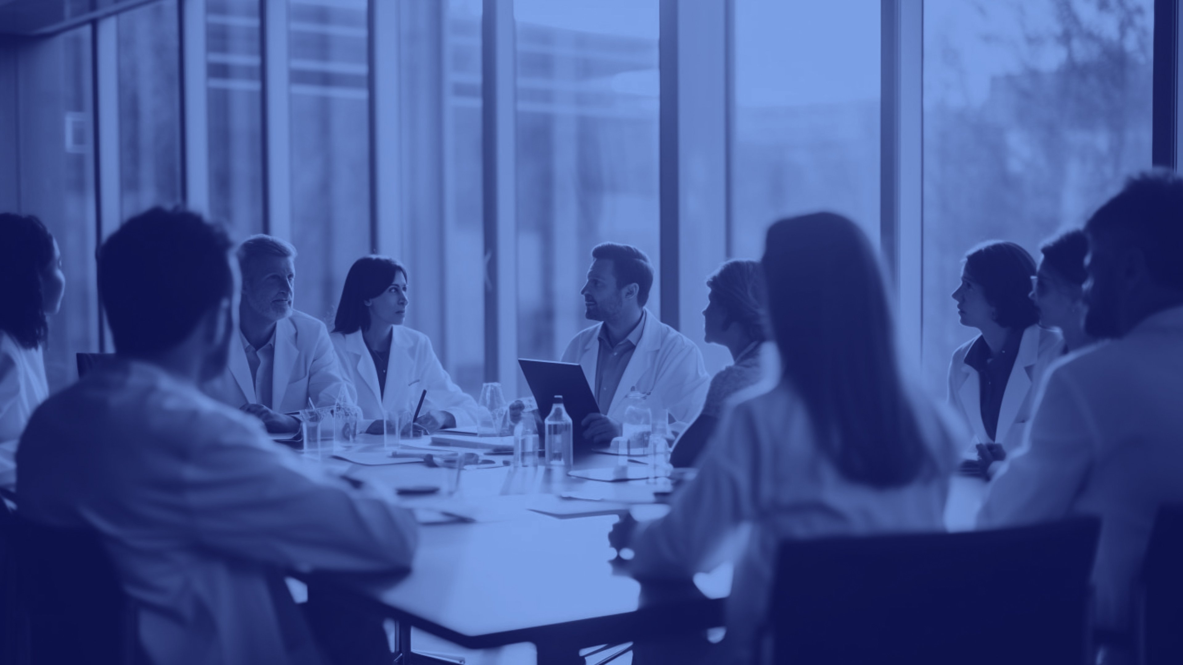 A team of professionals in lab coats seated around a boardroom table in front of large windows, actively discussing and planning. This image represents strategic collaboration and aligns with the messaging about delivering game-changing breakthroughs through planning methodologies like Hoshin Kanri and Agile.