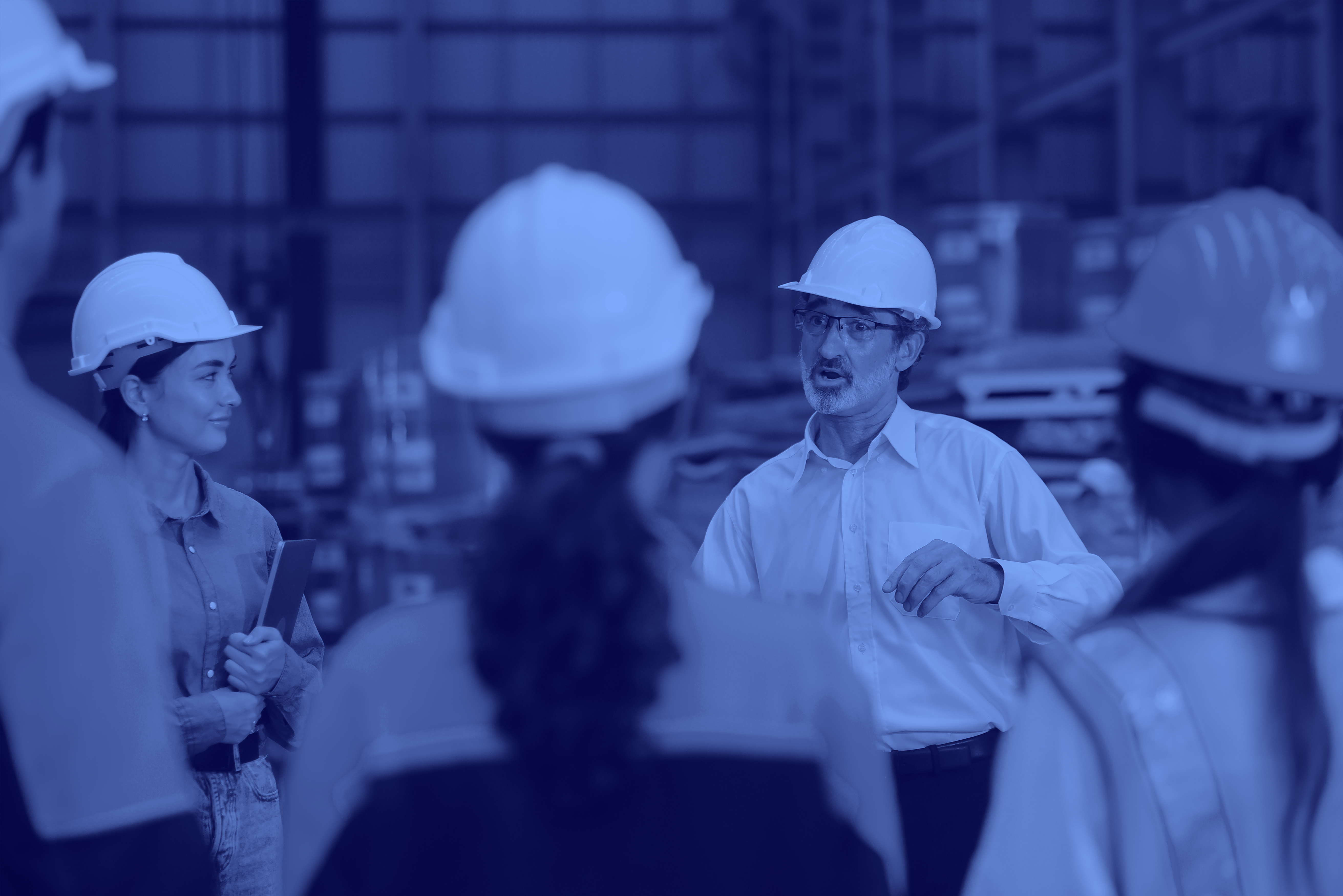 A group of professionals wearing white hard hats and safety gear, gathered in a warehouse or industrial setting, listening to a man who appears to be explaining. This image reflects the theme of delivering consistent year-on-year productivity improvements in operational environments.